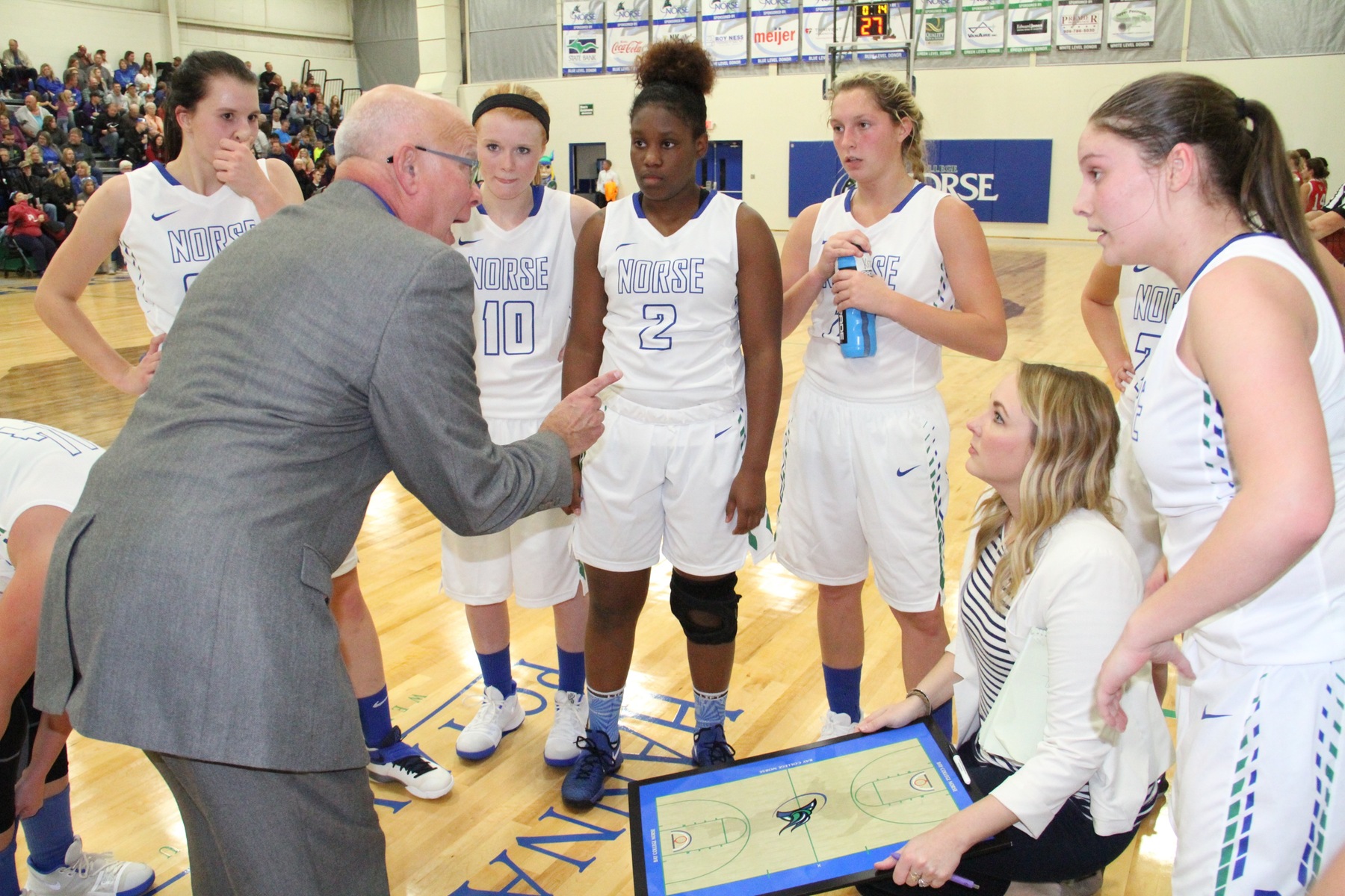 The team surrounds Rae Drake as he points, Kaite Polma holds the dry erase board, kneeling in the middle of the huddle